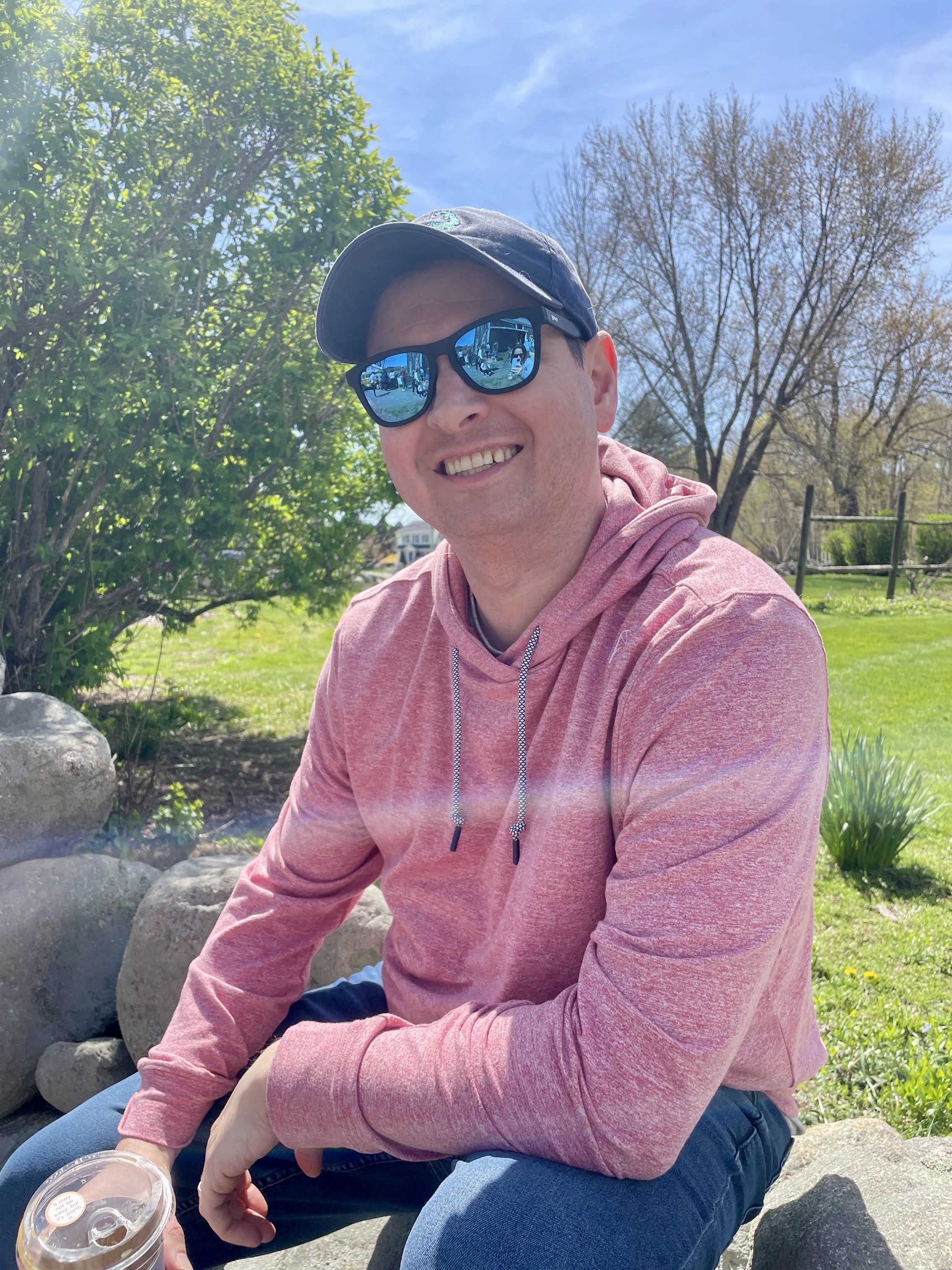 A white man with dark hair, wearing a red hoodie with sunglasses and a baseball cap, holding an iced coffee on a sunny day.